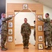 A U.S. Army military ceremony is taking place in an indoor facility, featuring a sword arch ritual. Two soldiers on either side of a decorative wooden arch are crossing sabers overhead while a central soldier stands at attention beneath the arch. The wooden arch displays multiple NCO rank insignia plaques and a unit crest at the base.
