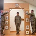 A U.S. Army NCO induction or promotion ceremony is taking place indoors. Two soldiers in OCP (Operational Camouflage Pattern) uniforms hold crossed sabers over a third soldier standing beneath a ceremonial wooden arch displaying NCO rank insignia. A unit crest featuring a rearing horse (likely Pennsylvania National Guard) is visible in a framed display at the base of the arch.