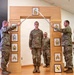 A U.S. Army promotion or induction ceremony is taking place indoors, featuring a soldier standing beneath a ceremonial arch while two soldiers cross sabers over his head. The wooden arch displays multiple NCO rank insignia plaques from Specialist through Sergeant Major. A framed unit crest (featuring a horse, consistent with a cavalry unit) is visible on the floor at the base of the arch.