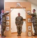 Three U.S. Army soldiers are participating in a formal NCO induction or promotion ceremony. Two soldiers on either side hold crossed sabers/swords over a third soldier standing at attention beneath a ceremonial wooden arch. The arch is decorated with NCO rank insignia plaques, and a unit crest is displayed on the floor in front.