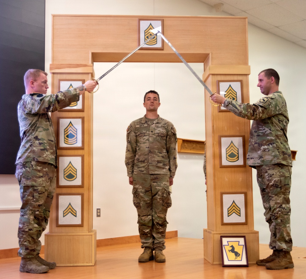 Three U.S. Army soldiers are participating in a formal NCO induction ceremony. Two soldiers on either side hold crossed sabers over a third soldier standing at attention beneath a ceremonial arch. The arch displays NCO rank insignia from Private through Sergeant Major, and a unit crest is visible at the base.