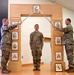 Three U.S. Army soldiers are participating in a formal NCO induction ceremony. Two soldiers on either side hold crossed sabers over a third soldier standing at attention beneath a ceremonial arch. The arch displays NCO rank insignia from Private through Sergeant Major, and a unit crest is visible at the base.