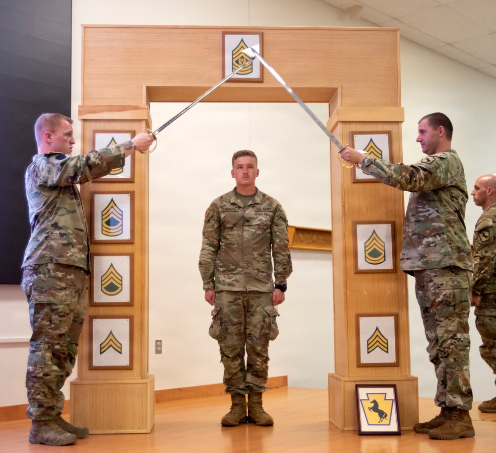 A U.S. Army promotion or graduation ceremony is taking place indoors, featuring a ceremonial arch with NCO rank insignia plaques. Two soldiers in OCP (Operational Camouflage Pattern) uniforms cross sabers/swords over a third soldier standing at attention beneath the arch. The ceremony appears to be an NCO induction or promotion ceremony, likely associated with the Pennsylvania National Guard based on visible unit insignia.