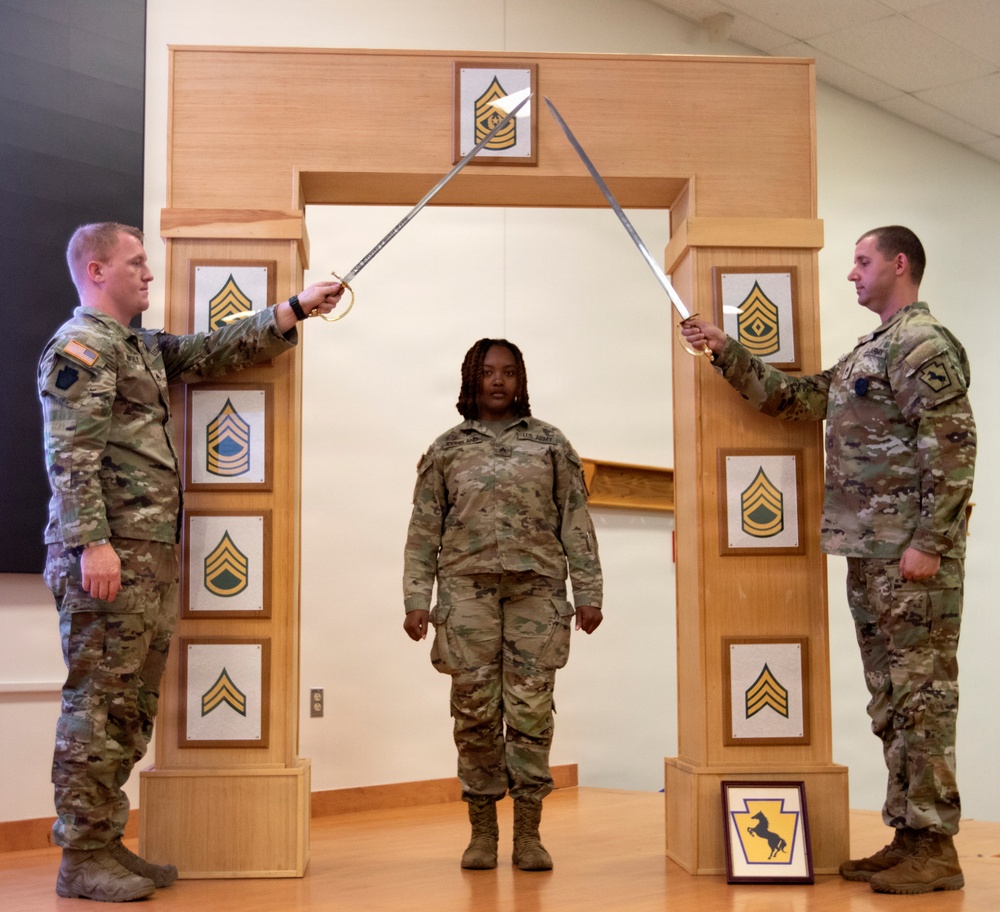 Three U.S. Army soldiers are participating in a formal NCO induction or promotion ceremony. Two soldiers holding ceremonial sabers form an arch over a third soldier standing at attention beneath a wooden arch structure displaying NCO rank insignia plaques. The ceremony appears to take place indoors in a military facility.