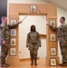 Three U.S. Army soldiers are participating in a formal NCO induction or promotion ceremony. Two soldiers holding ceremonial sabers form an arch over a third soldier standing at attention beneath a wooden arch structure displaying NCO rank insignia plaques. The ceremony appears to take place indoors in a military facility.