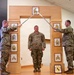 Three U.S. Army soldiers are participating in a formal NCO induction or promotion ceremony. Two soldiers on either side are crossing sabers/swords over a ceremonial arch while a third soldier stands at attention beneath the arch. The wooden arch displays various NCO rank insignia plaques and a unit crest, indicating this is an official Non-Commissioned Officer Corps ceremony.