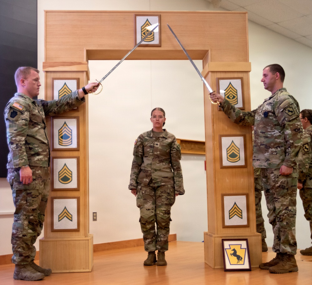 A U.S. Army promotion or induction ceremony is taking place indoors, featuring a ceremonial arch with NCO rank insignia plaques. Two soldiers holding sabers form an arch over a third soldier standing at attention in the center. The arch and framed insignia suggest this is a formal Non-Commissioned Officer (NCO) induction or promotion ceremony.