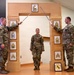 A U.S. Army promotion or induction ceremony is taking place indoors, featuring a ceremonial arch with NCO rank insignia plaques. Two soldiers holding sabers form an arch over a third soldier standing at attention in the center. The arch and framed insignia suggest this is a formal Non-Commissioned Officer (NCO) induction or promotion ceremony.