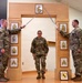 Three U.S. Army soldiers are posed in front of a decorative wooden arch displaying NCO rank insignia plaques, with two soldiers crossing sabers overhead in a ceremonial arch formation. A female soldier stands centered beneath the crossed sabers, suggesting this is a promotion or induction ceremony. A framed unit crest (appearing to be Pennsylvania National Guard) is visible on the floor at the base of the arch.