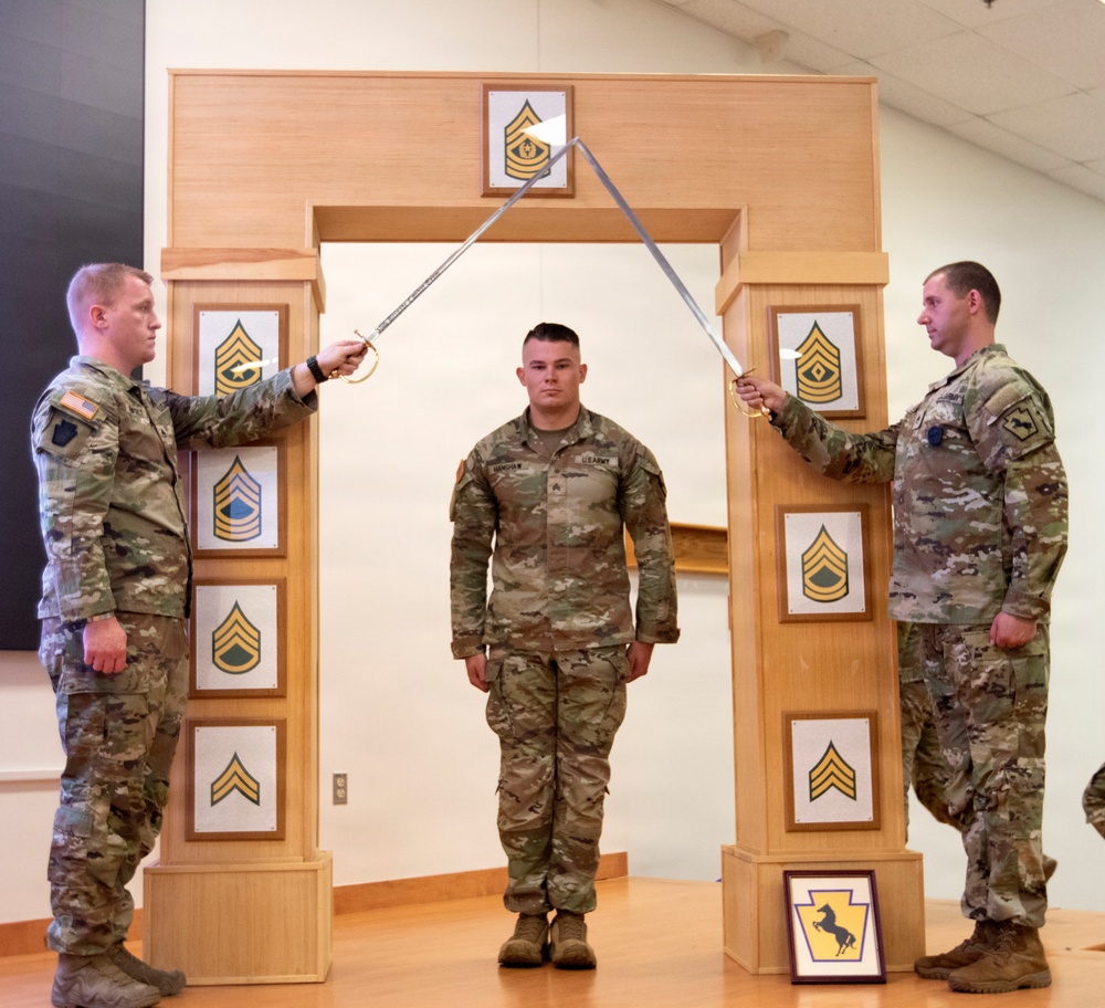 Three U.S. Army soldiers are participating in a formal NCO induction ceremony. Two soldiers on either side hold crossed sabers over a third soldier standing at attention beneath a ceremonial arch. The arch displays various NCO rank insignia plaques and a unit crest at the base.