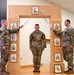 Three U.S. Army soldiers are participating in a formal NCO induction ceremony. Two soldiers on either side hold crossed sabers over a third soldier standing at attention beneath a ceremonial arch. The arch displays various NCO rank insignia plaques and a unit crest at the base.