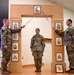 Three U.S. Army soldiers are participating in a formal NCO induction or promotion ceremony. Two soldiers on either side hold crossed sabers over a third soldier standing at attention beneath a ceremonial arch displaying NCO rank insignia plaques. The arch features enlisted rank chevrons from Corporal through Sergeant Major, and a unit crest is visible at the base.