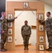 Three U.S. Army soldiers are participating in a formal NCO induction ceremony. Two soldiers holding ceremonial sabers form an arch over a third soldier standing at attention beneath a decorative wooden arch displaying NCO rank insignia plaques. A framed unit crest is visible on the floor in the foreground.
