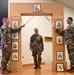 Three U.S. Army soldiers are participating in a formal NCO induction ceremony. Two soldiers holding sabers form an arch over a third soldier standing beneath a decorative wooden gate displaying NCO rank insignia plaques. A unit crest (rearing horse on yellow background) is displayed in a frame on the floor.