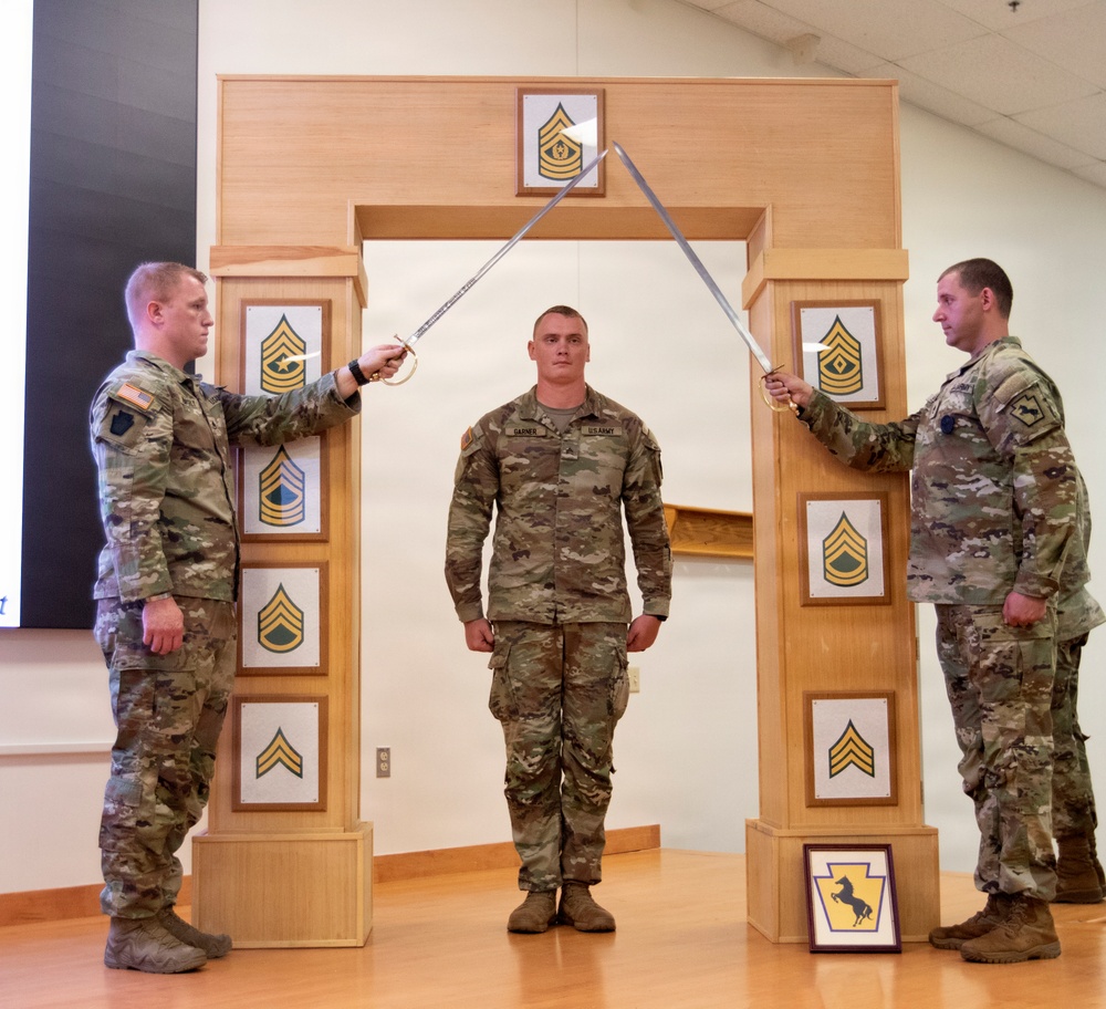 Three U.S. Army soldiers are participating in a formal NCO induction ceremony. Two soldiers on either side hold crossed sabers over a central soldier standing beneath a ceremonial arch. The arch displays various NCO rank insignia plaques, and a unit crest is visible on the floor in front of the structure.