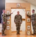 Three U.S. Army soldiers are participating in a formal NCO induction ceremony. Two soldiers on either side hold crossed sabers over a central soldier standing beneath a ceremonial arch. The arch displays various NCO rank insignia plaques, and a unit crest is visible on the floor in front of the structure.