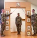 Three U.S. Army soldiers are participating in a formal military promotion or induction ceremony. Two soldiers on either side are crossing sabers/swords over a decorative wooden arch displaying NCO rank insignia plaques, while a third soldier stands at attention beneath the arch. A unit crest/patch is displayed at the base of the arch.