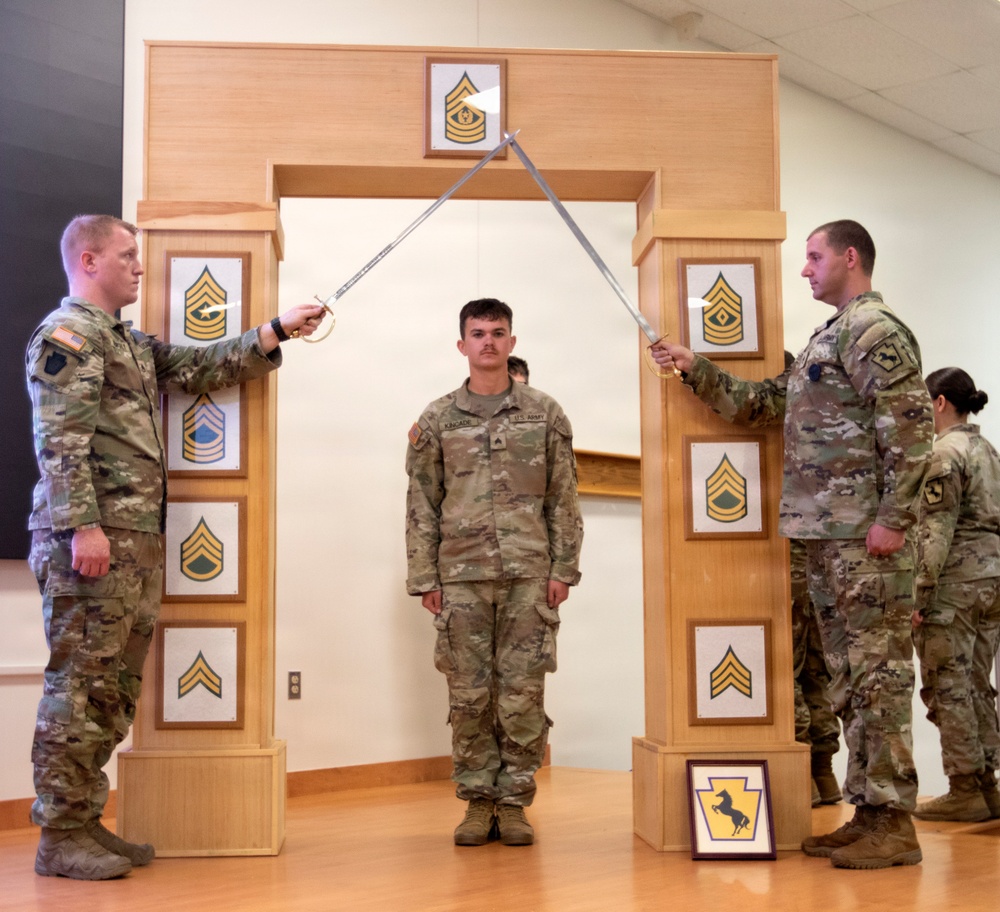A U.S. Army promotion or induction ceremony is taking place indoors, where a soldier stands beneath crossed sabers held by two NCOs flanking a decorative wooden arch. The arch displays multiple NCO rank insignia plaques progressing from Corporal to Sergeant Major, with a Command Sergeant Major insignia at the top. A framed unit patch (featuring a horse, consistent with a cavalry or armored unit) is visible on the floor at the base of the arch.