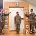 A U.S. Army promotion or induction ceremony is taking place indoors, where a soldier stands beneath crossed sabers held by two NCOs flanking a decorative wooden arch. The arch displays multiple NCO rank insignia plaques progressing from Corporal to Sergeant Major, with a Command Sergeant Major insignia at the top. A framed unit patch (featuring a horse, consistent with a cavalry or armored unit) is visible on the floor at the base of the arch.
