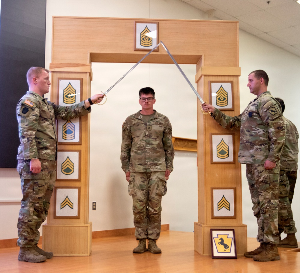 Three U.S. Army soldiers are participating in a formal NCO induction ceremony. Two soldiers on either side are crossing sabers/swords over a central soldier standing beneath a decorative wooden arch displaying NCO rank insignia progression. A framed unit crest (appearing to be the Pennsylvania National Guard's 28th Infantry Division) is visible at the base of the arch.