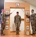 Three U.S. Army soldiers are participating in a formal NCO induction ceremony. Two soldiers on either side are crossing sabers/swords over a central soldier standing beneath a decorative wooden arch displaying NCO rank insignia progression. A framed unit crest (appearing to be the Pennsylvania National Guard's 28th Infantry Division) is visible at the base of the arch.