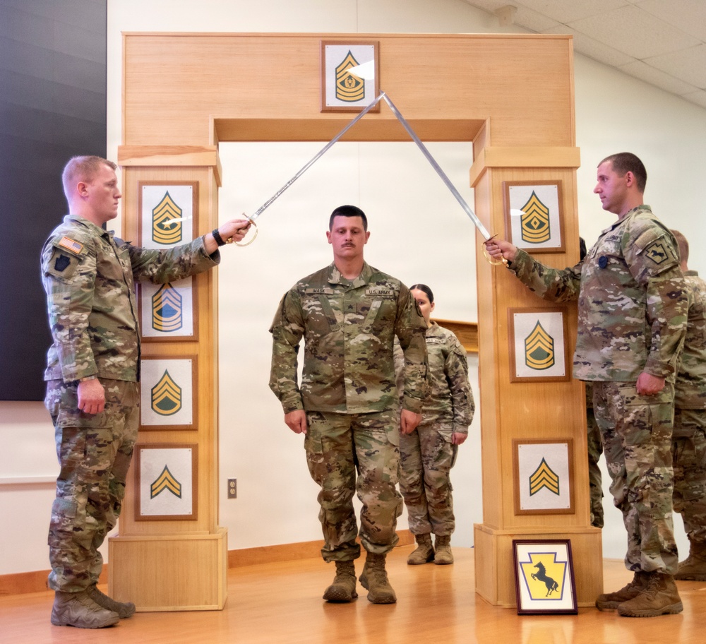 A U.S. Army promotion or induction ceremony is taking place indoors, featuring a soldier walking through an arch while two soldiers cross sabers overhead. The wooden arch displays multiple NCO rank insignia plaques ranging from Corporal to Sergeant Major. A framed unit patch, appearing to be the Pennsylvania Army National Guard (28th Infantry Division), is visible on the floor.