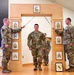 A U.S. Army promotion or induction ceremony is taking place indoors, featuring a soldier walking through an arch while two soldiers cross sabers overhead. The wooden arch displays multiple NCO rank insignia plaques ranging from Corporal to Sergeant Major. A framed unit patch, appearing to be the Pennsylvania Army National Guard (28th Infantry Division), is visible on the floor.