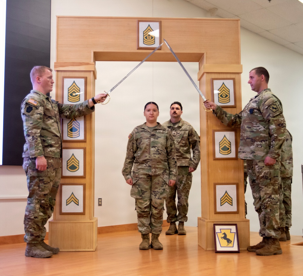 Four U.S. Army soldiers are participating in a formal NCO induction or promotion ceremony. Two soldiers on the flanks are crossing sabers/swords over an arch structure, while two soldiers stand beneath the arch at attention. The wooden arch displays NCO rank insignia panels from Specialist through Sergeant Major, with a Pennsylvania National Guard unit patch visible at the base.