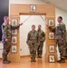 Four U.S. Army soldiers are participating in a formal NCO induction or promotion ceremony. Two soldiers on the flanks are crossing sabers/swords over an arch structure, while two soldiers stand beneath the arch at attention. The wooden arch displays NCO rank insignia panels from Specialist through Sergeant Major, with a Pennsylvania National Guard unit patch visible at the base.