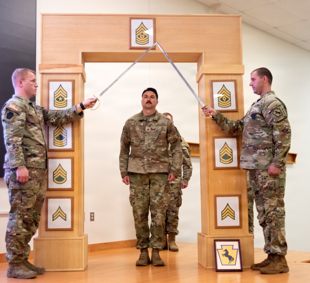 Three U.S. Army soldiers are participating in a formal military promotion or induction ceremony. Two soldiers on either side are crossing sabers/swords over a central soldier standing beneath a decorative wooden arch displaying NCO rank insignia plaques. A framed unit crest is visible on the floor in front of the arch.