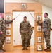 Three U.S. Army soldiers are participating in a formal military promotion or induction ceremony. Two soldiers on either side are crossing sabers/swords over a central soldier standing beneath a decorative wooden arch displaying NCO rank insignia plaques. A framed unit crest is visible on the floor in front of the arch.