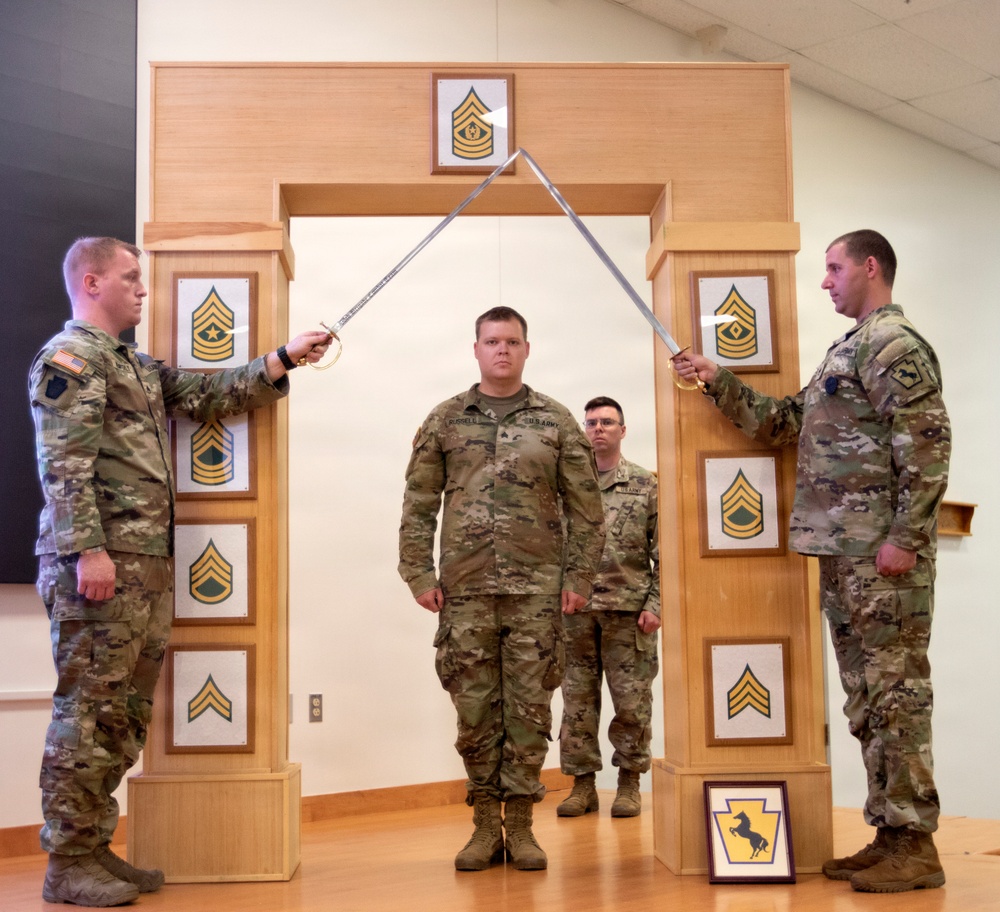 A U.S. Army promotion or induction ceremony is taking place indoors, featuring a ceremonial arch made of wood displaying NCO rank insignia plaques. Two soldiers on either side are crossing sabers/swords over a soldier standing beneath the arch, a traditional military ceremony gesture. A fourth soldier stands in the background observing the proceedings.