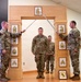 A U.S. Army promotion or induction ceremony is taking place indoors, featuring a ceremonial arch made of wood displaying NCO rank insignia plaques. Two soldiers on either side are crossing sabers/swords over a soldier standing beneath the arch, a traditional military ceremony gesture. A fourth soldier stands in the background observing the proceedings.