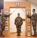 Three U.S. Army soldiers are participating in a formal NCO induction or promotion ceremony. Two soldiers on either side are crossing sabers/swords over a third soldier standing at attention beneath a decorative wooden arch displaying NCO rank insignia. A unit crest (appears to be 28th Infantry Division, Pennsylvania National Guard) is displayed at the base of the arch.