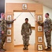 Three U.S. Army soldiers are participating in a formal military promotion ceremony. Two soldiers on either side are crossing sabers over a third soldier standing beneath a decorative wooden arch displaying NCO rank insignia plaques. This is a traditional NCO induction or promotion arch ceremony.