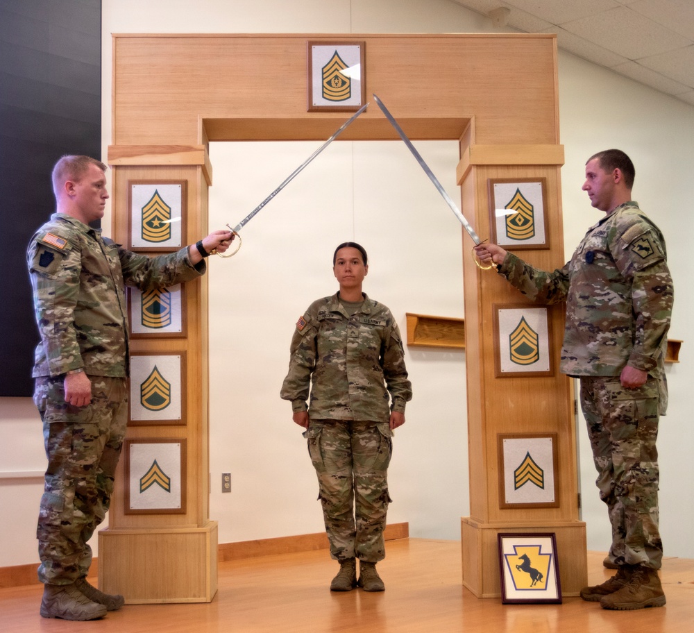 Three U.S. Army soldiers are participating in a formal military promotion or induction ceremony. Two soldiers holding ceremonial sabers form an arch over a third soldier standing at attention beneath a wooden arch structure displaying NCO rank insignia plaques. The arch and surrounding plaques represent the progression of Non-Commissioned Officer ranks from Private through Sergeant Major.