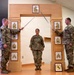 Three U.S. Army soldiers are participating in a formal military promotion or induction ceremony. Two soldiers holding ceremonial sabers form an arch over a third soldier standing at attention beneath a wooden arch structure displaying NCO rank insignia plaques. The arch and surrounding plaques represent the progression of Non-Commissioned Officer ranks from Private through Sergeant Major.