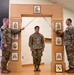 Three U.S. Army soldiers are participating in a formal military ceremony, likely an NCO induction or promotion ceremony. Two soldiers holding sabers form an arch over a third soldier standing at attention beneath a decorative wooden arch displaying NCO rank insignia plaques. A unit crest (appears to be 28th Infantry Division, Pennsylvania National Guard) is displayed in a frame on the floor.