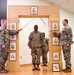A U.S. Army promotion or induction ceremony is taking place indoors, featuring a ceremonial arch decorated with NCO rank insignia plaques. Two soldiers in OCP (Operational Camouflage Pattern) uniforms are crossing sabers over a soldier standing beneath the arch, a traditional military ceremony. A framed unit crest (appearing to be the 1st Cavalry Division or similar horse insignia) is visible at the base of the arch.
