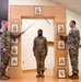 Three U.S. Army soldiers are participating in a formal NCO induction or promotion ceremony. Two soldiers on either side hold crossed sabers over a third soldier standing beneath a decorative wooden arch displaying NCO rank insignia plaques. A unit crest (appears to be 1st Cavalry Division or similar) is displayed in a frame at the base of the arch.