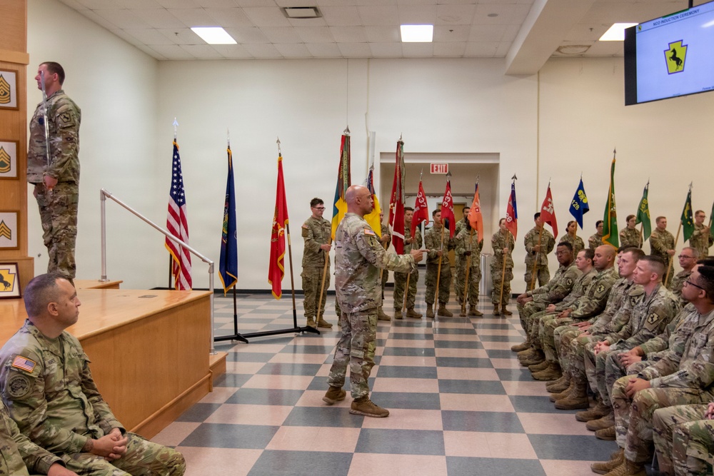 A formal U.S. Army military ceremony is taking place indoors, likely a unit induction or change of command ceremony as indicated by the screen in the upper right displaying 'NCO Induction Ceremony' with a horse insignia. Soldiers in Army Combat Uniforms (ACU/OCP pattern) are seated in rows facing a central figure holding a guidon or ceremonial staff, with multiple unit colors and flags displayed. A senior NCO stands at a podium on a raised platform to the left.