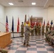 A formal U.S. Army military ceremony is taking place indoors, likely a unit induction or change of command ceremony as indicated by the screen in the upper right displaying 'NCO Induction Ceremony' with a horse insignia. Soldiers in Army Combat Uniforms (ACU/OCP pattern) are seated in rows facing a central figure holding a guidon or ceremonial staff, with multiple unit colors and flags displayed. A senior NCO stands at a podium on a raised platform to the left.