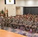 A military formation ceremony is taking place in an indoor facility, likely a gymnasium or assembly hall. A senior officer or NCO is addressing a large group of seated soldiers in Army Combat Uniforms (ACU/OCP pattern). A color guard with multiple unit flags is visible in the background, and a screen displaying what appears to be the 1st Cavalry Division patch is mounted on the wall.
