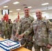 Two U.S. Army soldiers, identified by name tapes as KLINE and MELENDEZ, pose together while cutting a decorated military cake at what appears to be a formal unit ceremony or celebration. Sergeant Major Melendez, identifiable by his rank insignia, stands alongside Soldier Kline in a large indoor military facility. Numerous other soldiers in multicam uniforms stand in formation in the background, with red unit guidon flags visible.