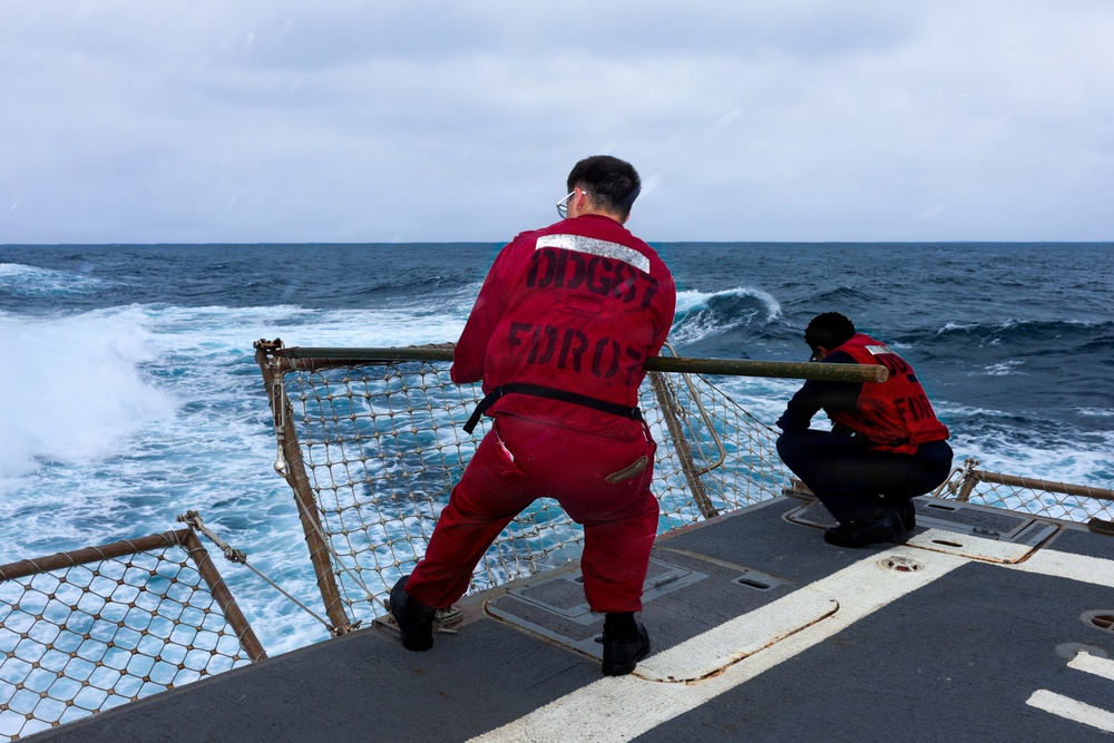 Two personnel in red coveralls and life vests stand at the stern railing of a military or coast guard vessel, observing rough open ocean waters. The individual in the foreground wears a full red jumpsuit with stenciled text on the back reading 'DCGST FORO7' or similar markings. The sea conditions appear moderately rough with visible wake and whitecaps under an overcast sky.