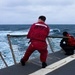 Two personnel in red coveralls and life vests stand at the stern railing of a military or coast guard vessel, observing rough open ocean waters. The individual in the foreground wears a full red jumpsuit with stenciled text on the back reading 'DCGST FORO7' or similar markings. The sea conditions appear moderately rough with visible wake and whitecaps under an overcast sky.