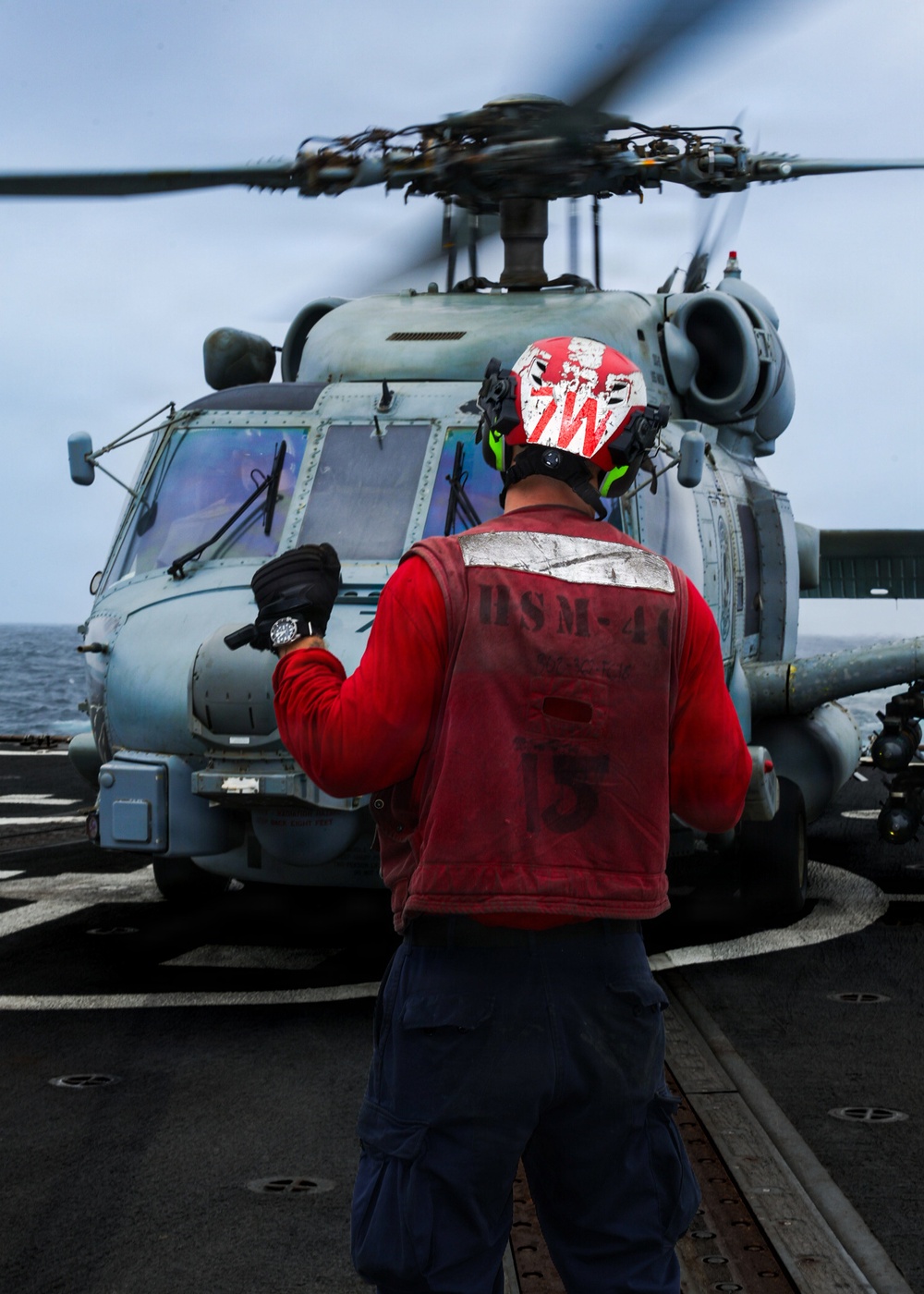 A U.S. Navy flight deck crew member wearing a red jersey and helmet is marshaling a military helicopter on a ship's flight deck at sea. The crew member's vest is marked 'HSM-41' identifying the Helicopter Maritime Strike Squadron 41. The helicopter, consistent with an MH-60R Seahawk, has its rotors spinning and is positioned on the deck with the ocean visible in the background.