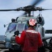 A U.S. Navy flight deck crew member wearing a red jersey and helmet is marshaling a military helicopter on a ship's flight deck at sea. The crew member's vest is marked 'HSM-41' identifying the Helicopter Maritime Strike Squadron 41. The helicopter, consistent with an MH-60R Seahawk, has its rotors spinning and is positioned on the deck with the ocean visible in the background.
