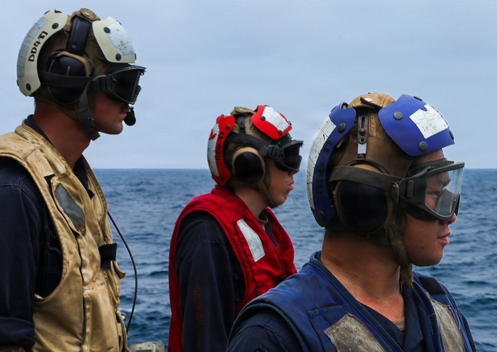 Three military personnel are standing on the deck of a naval vessel at sea, each wearing color-coded helmets and protective gear consistent with flight deck or damage control roles. The ocean is visible in the background under an overcast sky. Their different vest and helmet colors indicate distinct crew roles or assignments.
