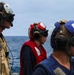 Three military personnel are standing on the deck of a naval vessel at sea, each wearing color-coded helmets and protective gear consistent with flight deck or damage control roles. The ocean is visible in the background under an overcast sky. Their different vest and helmet colors indicate distinct crew roles or assignments.
